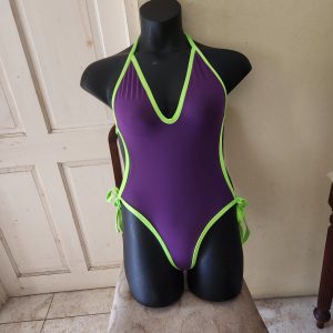 Purple one-piece swimsuit with neon green trim, displayed on a black mannequin seated on a cushioned chair, with a neutral background.